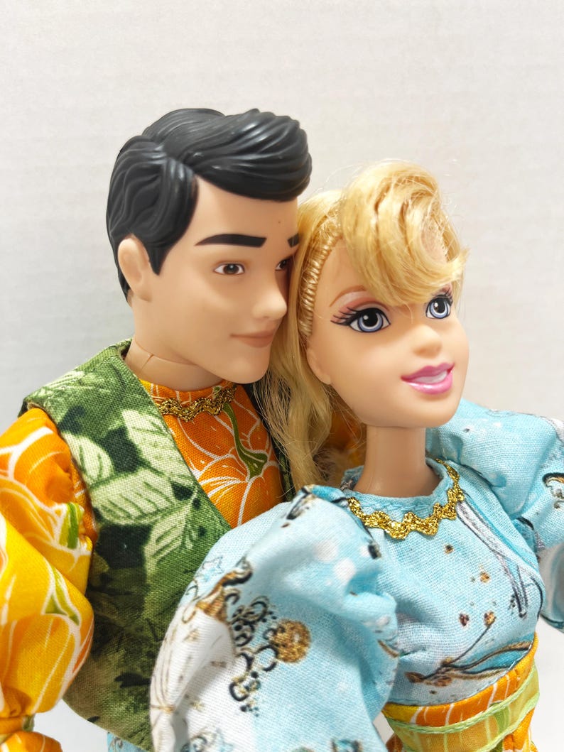 以下が含まれることがあります： 2体のファッションドール、男性と女性が寄り添っています。男性は黒髪で、緑とオレンジの模様のベストを着ています。女性はブロンドの髪で、金色のネックレスが付いた水色のドレスを着ています。