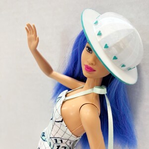 May include: A doll with vibrant blue hair wearing a white hat with small teal heart accents and a white dress featuring a black grid pattern, clock numbers including 11 and 12, and various graphic illustrations. The doll has bright pink lipstick.