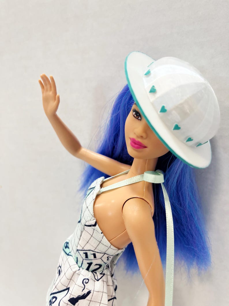 May include: A doll with vibrant blue hair wearing a white hat with small teal heart accents and a white dress featuring a black grid pattern, clock numbers including 11 and 12, and various graphic illustrations. The doll has bright pink lipstick.