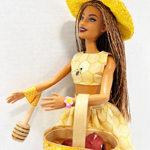 Puede incluir: Mu&ntilde;eca con un atuendo amarillo con estampado de panal, sombrero y pu&ntilde;os, sosteniendo una cesta con un objeto rojo y un cuchar&oacute;n de miel de madera. El atuendo presenta un gr&aacute;fico de abeja. La mu&ntilde;eca tiene largas trenzas marrones.