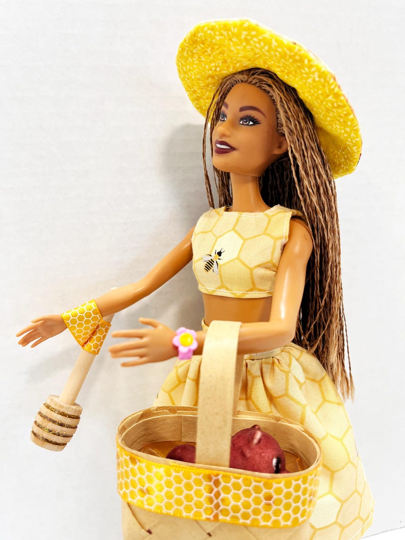 Puede incluir: Mu&ntilde;eca con un atuendo amarillo con estampado de panal, sombrero y pu&ntilde;os, sosteniendo una cesta con un objeto rojo y un cuchar&oacute;n de miel de madera. El atuendo presenta un gr&aacute;fico de abeja. La mu&ntilde;eca tiene largas trenzas marrones.