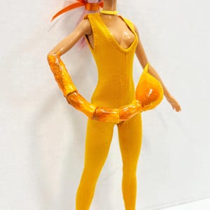 K&ouml;nnte beinhalten: Eine Puppe mit hellrosa Haar tr&auml;gt einen gelben &auml;rmellosen Jumpsuit, ein gelbes Halsband und violett get&ouml;nte Sonnenbrille. Die Puppe hat einen gl&auml;nzenden orangefarbenen Skorpionschwanz um die Taille und dazu passende orangefarbene High Heels.