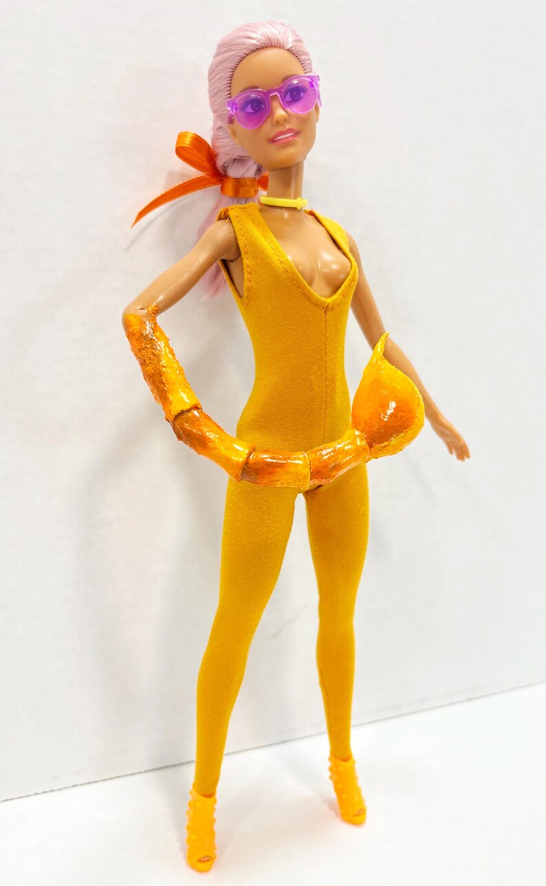 K&ouml;nnte beinhalten: Eine Puppe mit hellrosa Haar tr&auml;gt einen gelben &auml;rmellosen Jumpsuit, ein gelbes Halsband und violett get&ouml;nte Sonnenbrille. Die Puppe hat einen gl&auml;nzenden orangefarbenen Skorpionschwanz um die Taille und dazu passende orangefarbene High Heels.