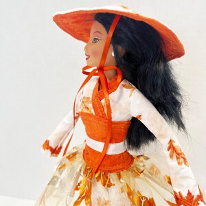 May include: A doll with long black hair wearing a wide-brimmed orange hat with a ribbon tie. The doll is dressed in a white long-sleeved top featuring an orange leaf pattern, a solid orange bodice, and a full skirt decorated with orange leaf motifs.
