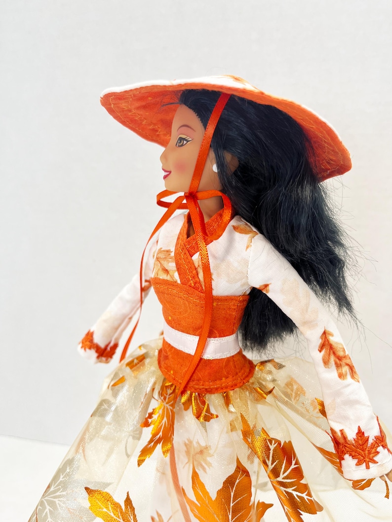 May include: A doll with long black hair wearing a wide-brimmed orange hat with a ribbon tie. The doll is dressed in a white long-sleeved top featuring an orange leaf pattern, a solid orange bodice, and a full skirt decorated with orange leaf motifs.