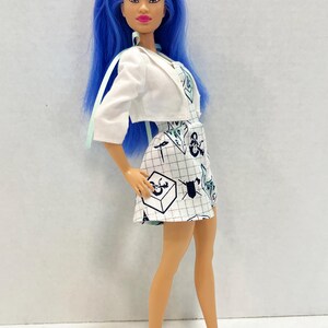 May include: A doll with long blue hair wearing a white cropped jacket over a white dress featuring a black grid pattern and various black and blue graphic symbols including shields and cubes. The doll wears simple white slip-on shoes on its feet.