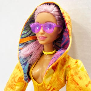 K&ouml;nnte beinhalten: Eine Puppe mit hellrosa Haar tr&auml;gt eine durchscheinende violette Sonnenbrille, ein gelbes Halsband und eine gelbe Kapuzenjacke. Die Jacke hat eine leuchtend gelbe Au&szlig;enseite mit Batik-Effekt und ein buntes Innenfutter mit blauen, violetten und orangefarbenen Mustern.