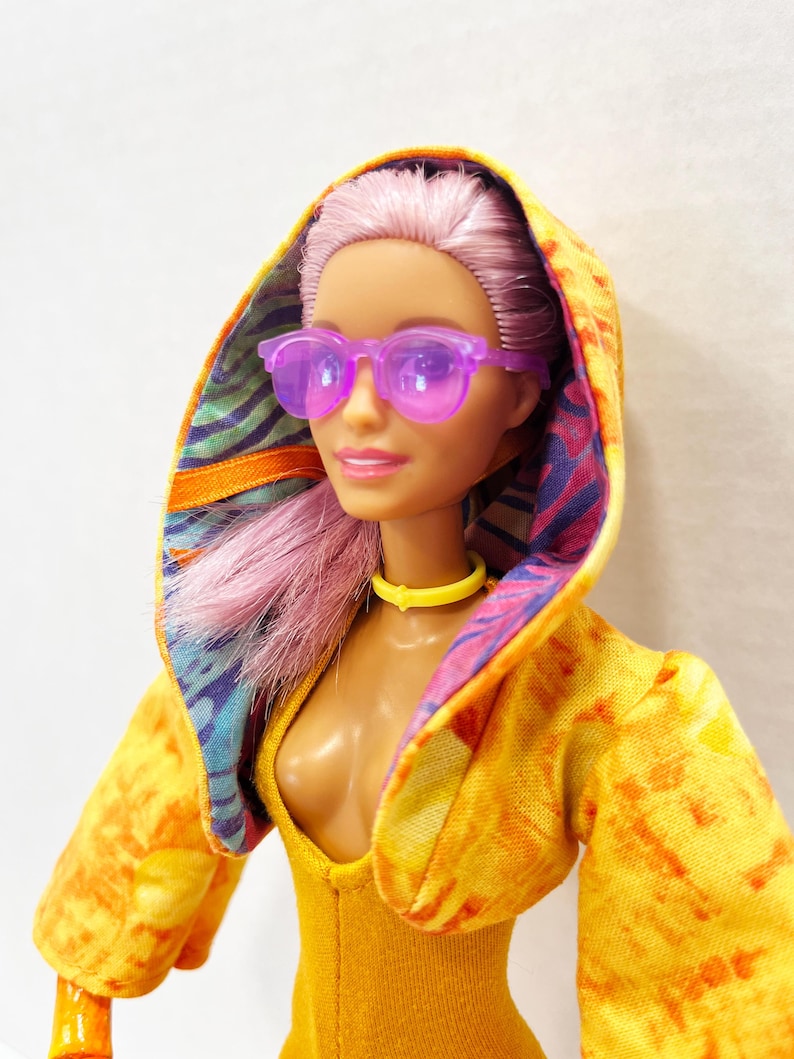 K&ouml;nnte beinhalten: Eine Puppe mit hellrosa Haar tr&auml;gt eine durchscheinende violette Sonnenbrille, ein gelbes Halsband und eine gelbe Kapuzenjacke. Die Jacke hat eine leuchtend gelbe Au&szlig;enseite mit Batik-Effekt und ein buntes Innenfutter mit blauen, violetten und orangefarbenen Mustern.