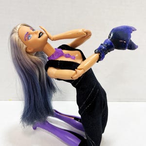 May include: A fashion doll with long, two-toned purple and silver hair, wearing a black dress with silver accents. The doll has purple eye makeup, black lipstick, and a purple glittery necklace. The doll is also wearing a dark blue glove.
