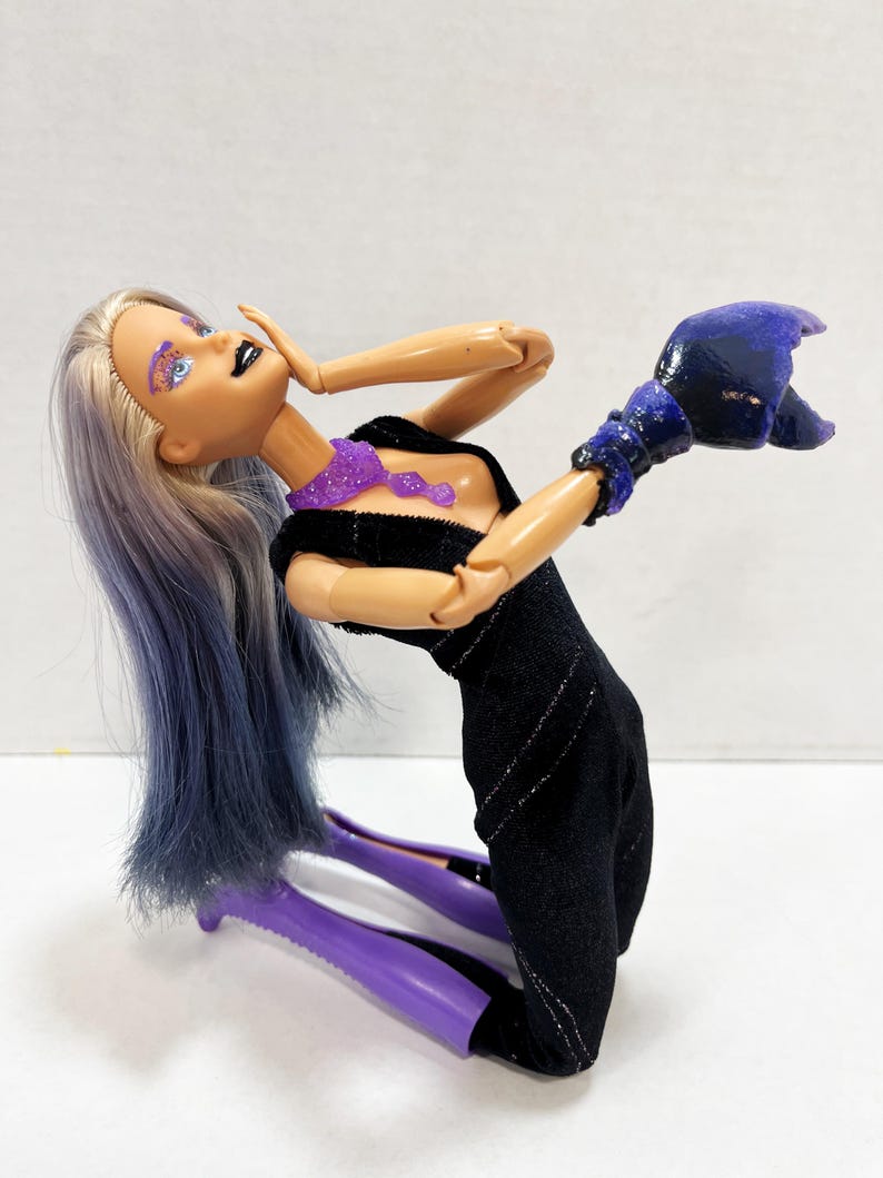 May include: A fashion doll with long, two-toned purple and silver hair, wearing a black dress with silver accents. The doll has purple eye makeup, black lipstick, and a purple glittery necklace. The doll is also wearing a dark blue glove.