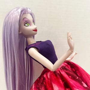 Pu&ograve; includere: Una bambola con lunghi capelli lisci color lavanda, pelle chiara e occhi verdi. La bambola indossa un corpetto viola scuro senza maniche con sottili cuciture rosa sullo scollo e un'ampia gonna arricciata con un motivo marmorizzato rosso e nero.