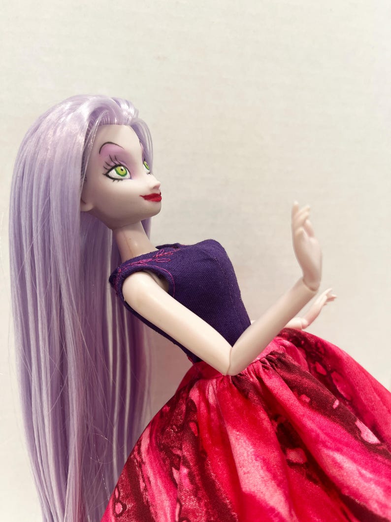 Pu&ograve; includere: Una bambola con lunghi capelli lisci color lavanda, pelle chiara e occhi verdi. La bambola indossa un corpetto viola scuro senza maniche con sottili cuciture rosa sullo scollo e un'ampia gonna arricciata con un motivo marmorizzato rosso e nero.