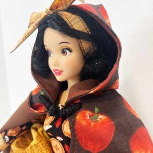 Op de afbeelding: Een pop met zwart haar draagt een bruine cape met capuchon en een rode appelprint. De pop heeft een bruine geruite hoofdband met een strik bovenop en een zwarte knevelsluiting bij de nek. Onder de cape is een zwarte jurk met taartprint zichtbaar.