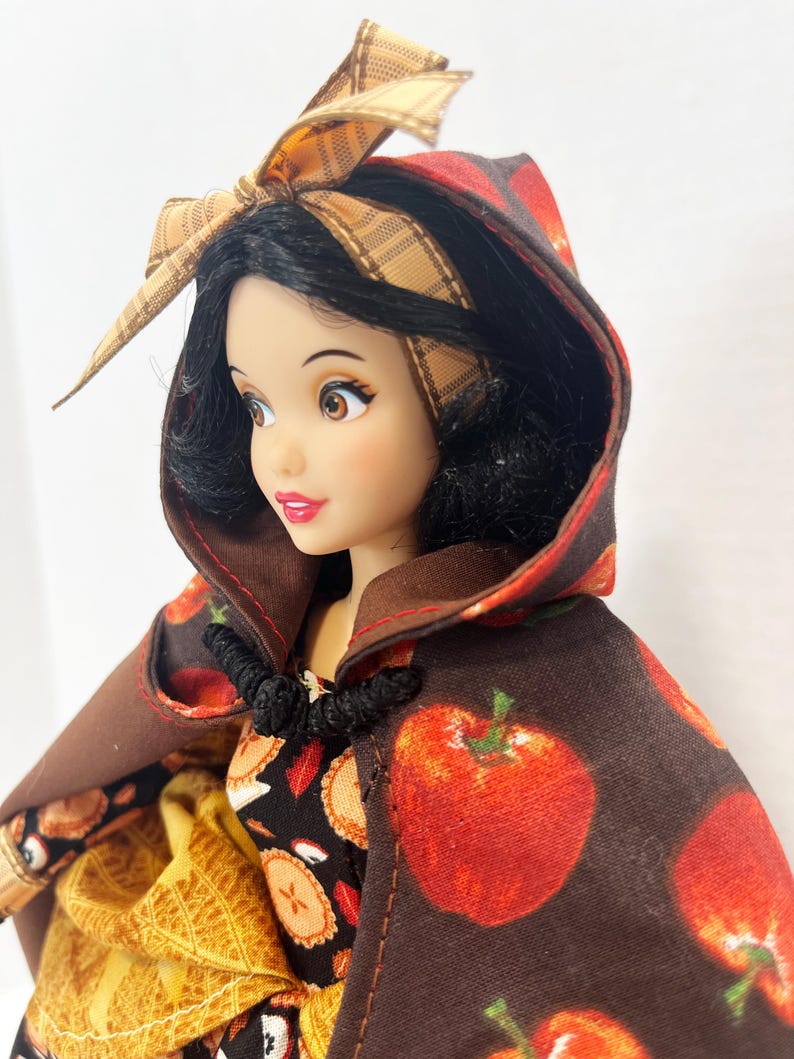 Op de afbeelding: Een pop met zwart haar draagt een bruine cape met capuchon en een rode appelprint. De pop heeft een bruine geruite hoofdband met een strik bovenop en een zwarte knevelsluiting bij de nek. Onder de cape is een zwarte jurk met taartprint zichtbaar.