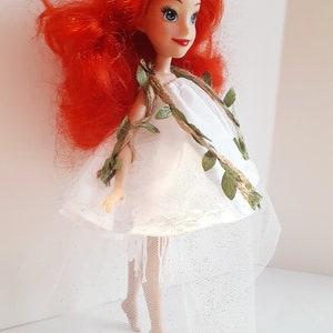 Puede incluir: Una mu&ntilde;eca de pelo rojo con un vestido blanco con una guirnalda de vid verde y una falda de tul blanca.