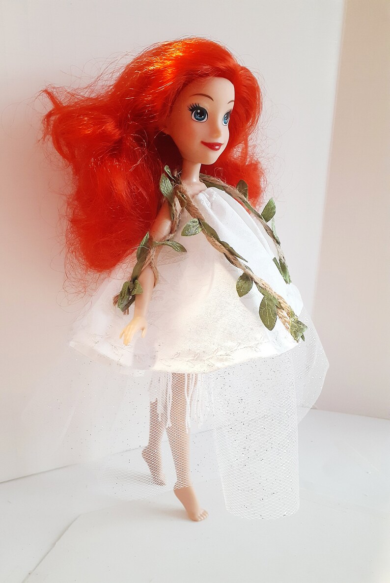 Puede incluir: Una mu&ntilde;eca de pelo rojo con un vestido blanco con una guirnalda de vid verde y una falda de tul blanca.