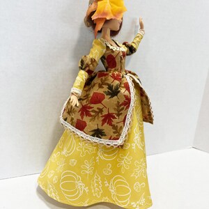 May include: A doll wearing a long yellow skirt with white line drawings of pumpkins and leaves, paired with a brown bodice featuring a red and orange autumn leaf print, white lace trim, and a straw hat adorned with a yellow flower and orange leaves.