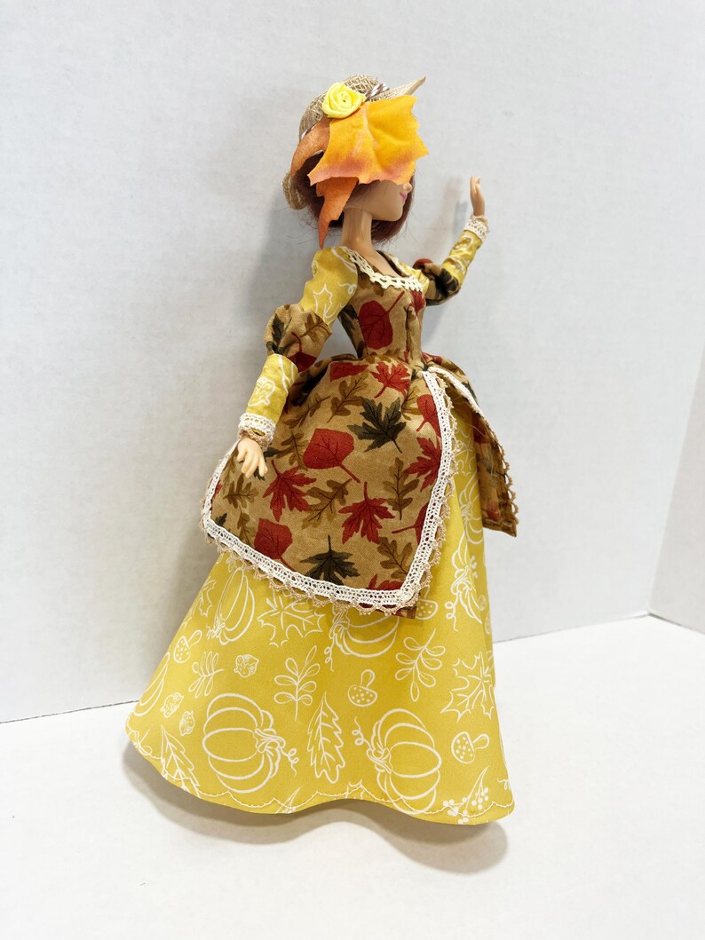 May include: A doll wearing a long yellow skirt with white line drawings of pumpkins and leaves, paired with a brown bodice featuring a red and orange autumn leaf print, white lace trim, and a straw hat adorned with a yellow flower and orange leaves.