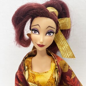 May include: A doll with dark reddish-purple hair styled in a high ponytail with a gold ribbon. The doll has light skin, purple eyeshadow, and dark red lipstick. It wears a yellow textured bodice with gold leaf accents and a dark red leaf-patterned shawl.