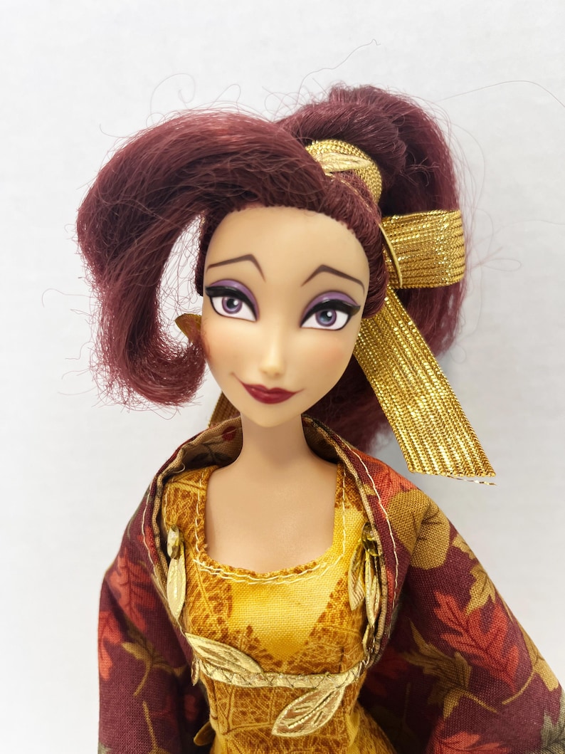 May include: A doll with dark reddish-purple hair styled in a high ponytail with a gold ribbon. The doll has light skin, purple eyeshadow, and dark red lipstick. It wears a yellow textured bodice with gold leaf accents and a dark red leaf-patterned shawl.