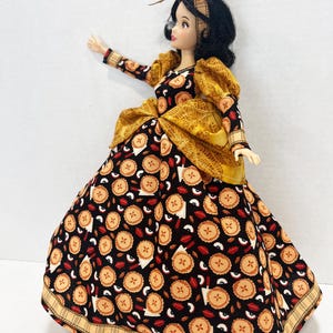 Op de afbeelding: Een pop met zwart haar draagt een hoofdband en een lange jurk met een zwarte stoffen basis met een herhalend patroon van kleine taartjes en appelschijfjes. De jurk heeft goudkleurige pofmouwen, een gouden tailleaccent en een geruite zoom.