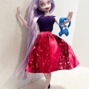 Pu&ograve; includere: Una bambola con lunghi capelli color lavanda, che indossa un top senza maniche viola scuro e una gonna rossa vivace con motivo maculato. La bambola ha occhi verdi, labbra rosse e tacchi viola, mentre tiene in mano una piccola creatura blu.