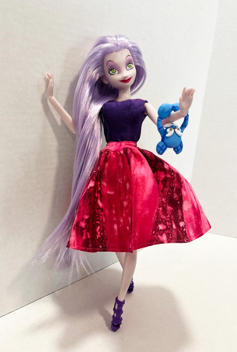 Pu&ograve; includere: Una bambola con lunghi capelli color lavanda, che indossa un top senza maniche viola scuro e una gonna rossa vivace con motivo maculato. La bambola ha occhi verdi, labbra rosse e tacchi viola, mentre tiene in mano una piccola creatura blu.