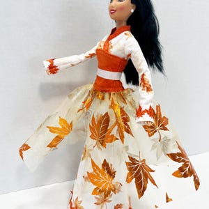 May include: A doll with long black hair wearing a white long-sleeved top with orange leaf patterns and a matching orange bodice. The skirt is a white floor-length gown featuring a sheer overlay decorated with metallic orange and brown maple leaf prints.