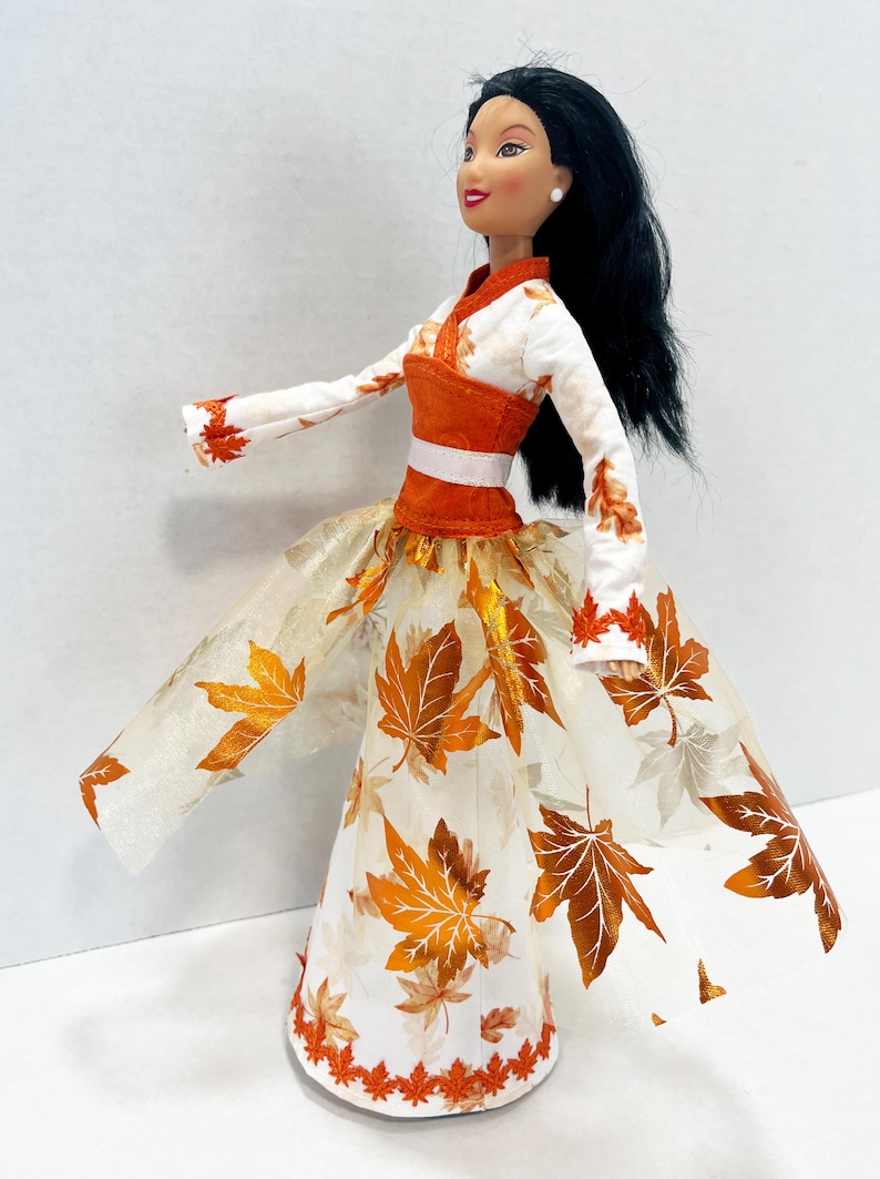 May include: A doll with long black hair wearing a white long-sleeved top with orange leaf patterns and a matching orange bodice. The skirt is a white floor-length gown featuring a sheer overlay decorated with metallic orange and brown maple leaf prints.