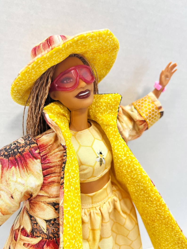 Puede incluir: Mu&ntilde;eca con un atuendo amarillo con dise&ntilde;o de abeja, que incluye sombrero, gafas de sol, top corto y falda. La mu&ntilde;eca tambi&eacute;n lleva un abrigo largo con estampado floral. La mu&ntilde;eca lleva una pulsera rosa.