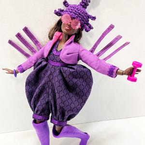 May include: A doll in a purple ensemble, including a hat and glasses, holding a pink dumbbell. The outfit features a jacket, dress, and boots. The hat is adorned with a decorative top with pink and green details. The doll is on a white background.