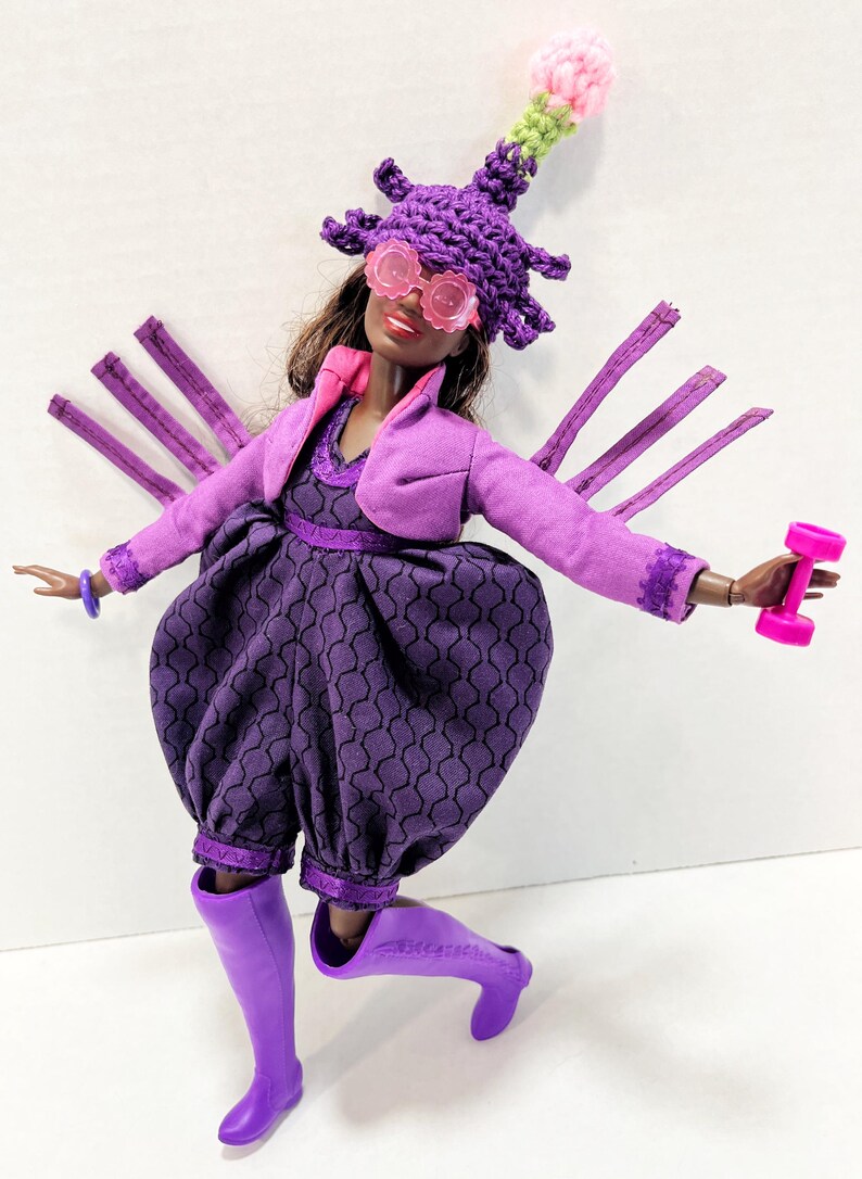 May include: A doll in a purple ensemble, including a hat and glasses, holding a pink dumbbell. The outfit features a jacket, dress, and boots. The hat is adorned with a decorative top with pink and green details. The doll is on a white background.
