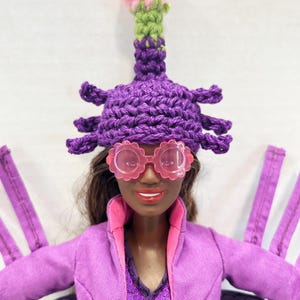 May include: A doll wearing a purple crocheted hat with a pink flower, pink glasses, and a purple jacket. The doll has brown hair and is wearing a purple dress. The hat has a unique design.