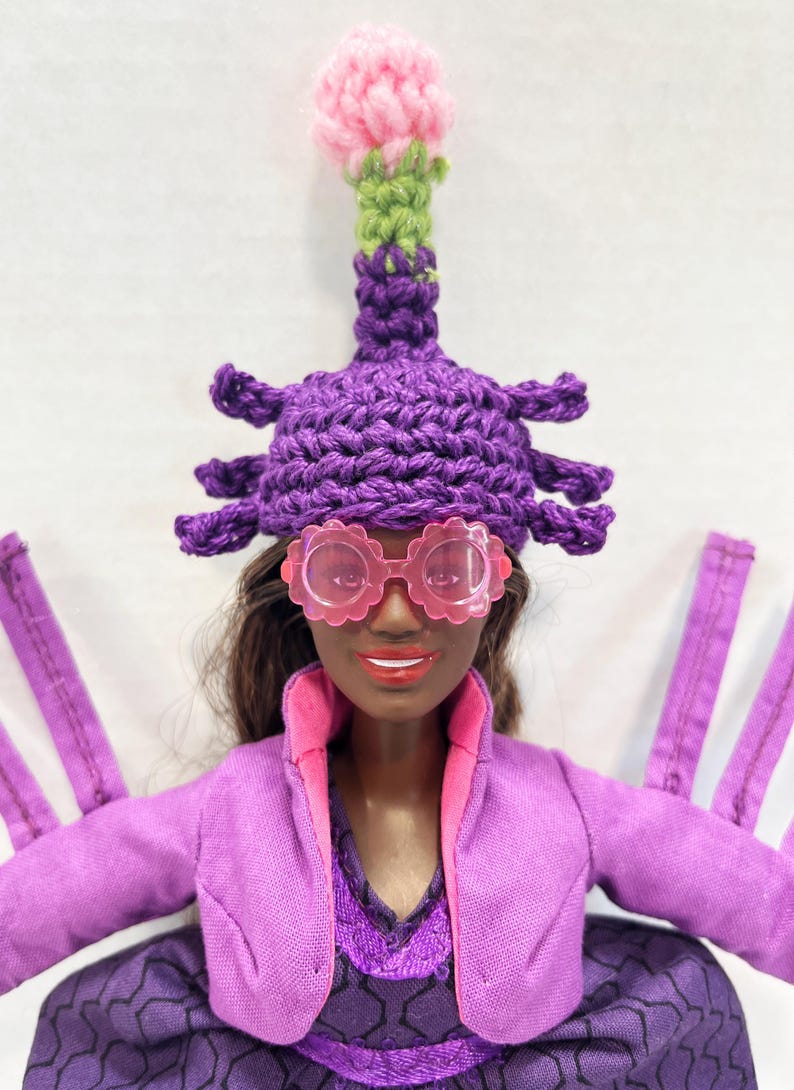 May include: A doll wearing a purple crocheted hat with a pink flower, pink glasses, and a purple jacket. The doll has brown hair and is wearing a purple dress. The hat has a unique design.