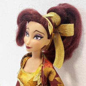 May include: A doll with long, dark red hair styled in a high ponytail with a gold metallic ribbon. The doll has light skin, purple eyeshadow, and dark red lipstick. It wears a yellow and maroon dress featuring a printed leaf pattern and gold trim.