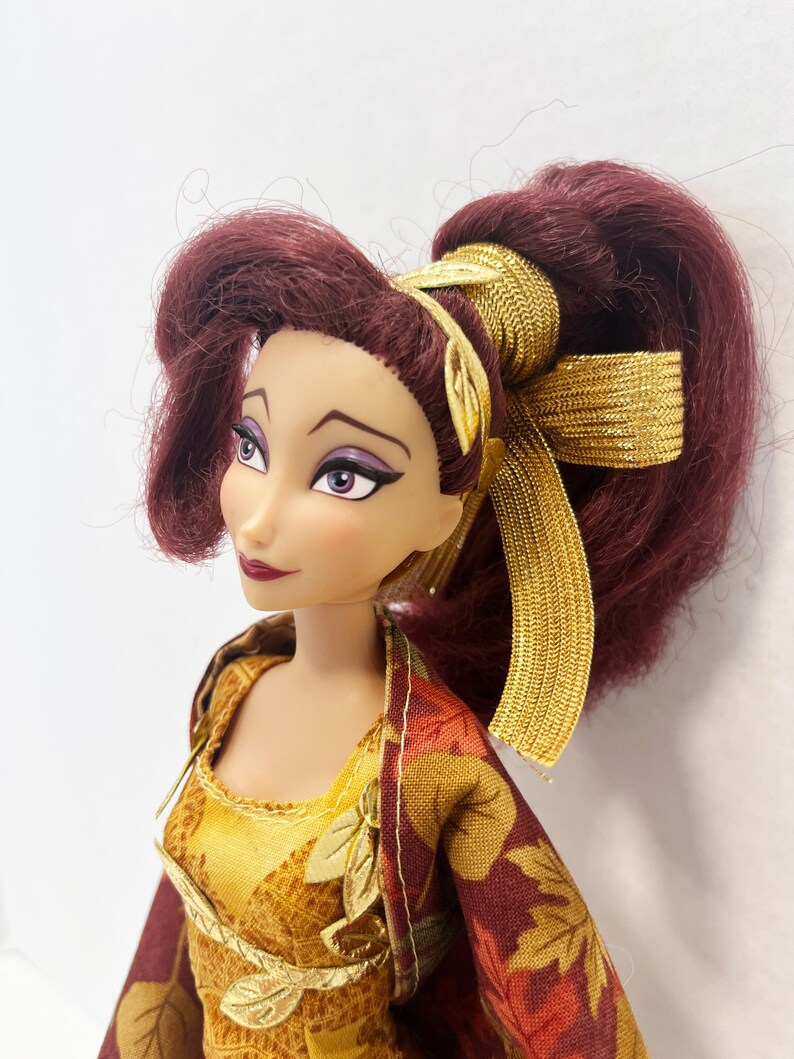 May include: A doll with long, dark red hair styled in a high ponytail with a gold metallic ribbon. The doll has light skin, purple eyeshadow, and dark red lipstick. It wears a yellow and maroon dress featuring a printed leaf pattern and gold trim.