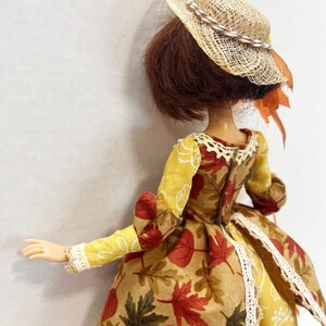 May include: A doll with short brown hair wearing a straw hat with a braided band and orange leaf accents. The doll is dressed in a long-sleeved gown featuring a yellow fabric with white line drawings and a tan fabric with a red and green leaf print.