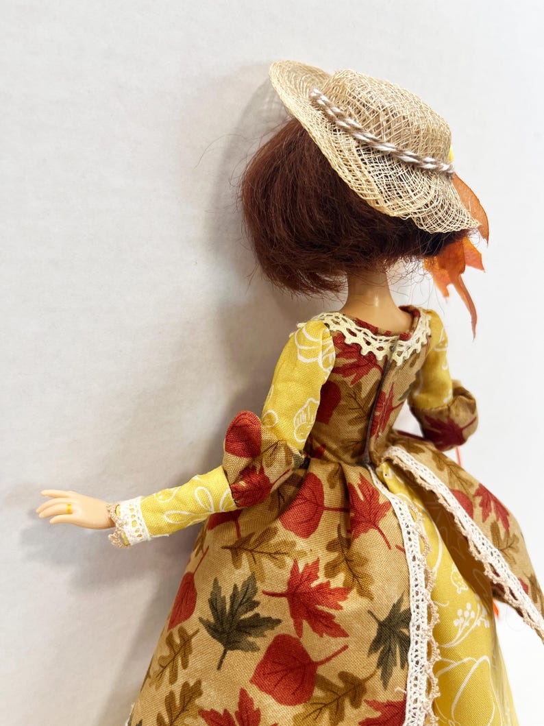 May include: A doll with short brown hair wearing a straw hat with a braided band and orange leaf accents. The doll is dressed in a long-sleeved gown featuring a yellow fabric with white line drawings and a tan fabric with a red and green leaf print.