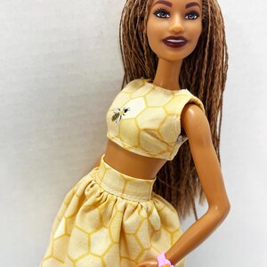 Puede incluir: Mu&ntilde;eca con un conjunto de top corto y falda amarillos con estampado de panal. El top presenta un peque&ntilde;o gr&aacute;fico de abeja. La mu&ntilde;eca tiene pelo largo casta&ntilde;o y una pulsera de flores rosa. El atuendo es un estilo veraniego y divertido.