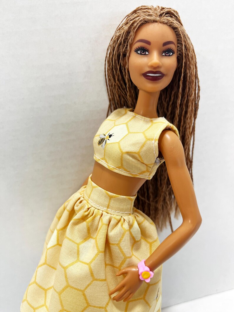 Puede incluir: Mu&ntilde;eca con un conjunto de top corto y falda amarillos con estampado de panal. El top presenta un peque&ntilde;o gr&aacute;fico de abeja. La mu&ntilde;eca tiene pelo largo casta&ntilde;o y una pulsera de flores rosa. El atuendo es un estilo veraniego y divertido.