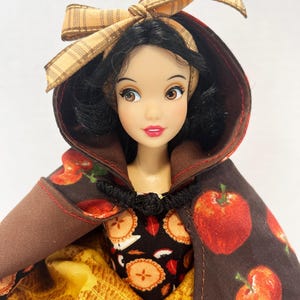 Op de afbeelding: Een pop met zwart haar draagt een bruine cape met capuchon, een rode appelprint en een zwarte knevelsluiting. De pop draagt een jurk met een zwart lijfje met een patroon van appeltaarten en een gele rok met een gestructureerd ruitpatroon.
