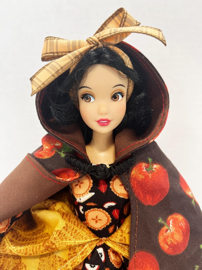 Op de afbeelding: Een pop met zwart haar draagt een bruine cape met capuchon, een rode appelprint en een zwarte knevelsluiting. De pop draagt een jurk met een zwart lijfje met een patroon van appeltaarten en een gele rok met een gestructureerd ruitpatroon.