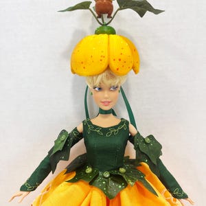 May include: A doll with blonde hair wearing a yellow flower-shaped hat topped with a small brown figure. The doll wears a dark green bodice with leaf-like sleeves and a long yellow skirt featuring white eyelet embroidery and green leaf accents.