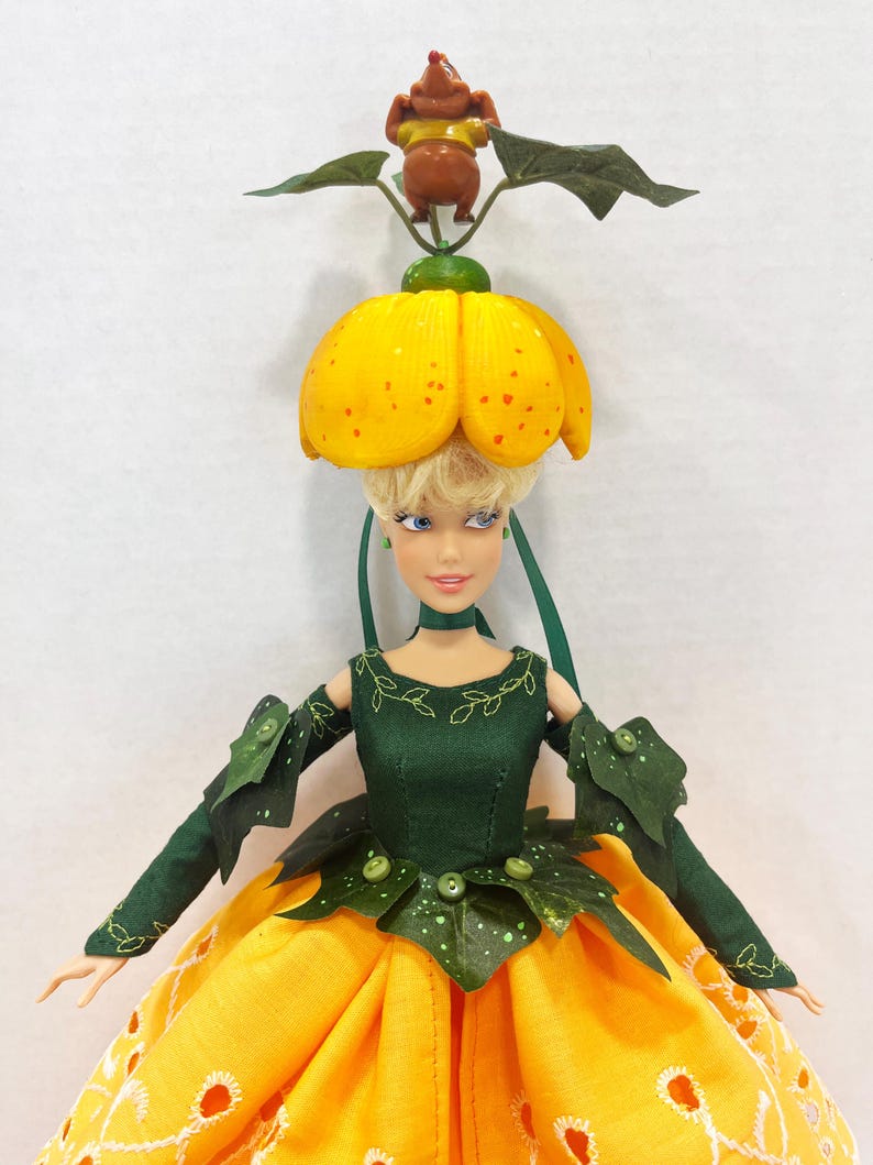 May include: A doll with blonde hair wearing a yellow flower-shaped hat topped with a small brown figure. The doll wears a dark green bodice with leaf-like sleeves and a long yellow skirt featuring white eyelet embroidery and green leaf accents.