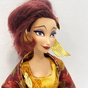 May include: A doll with reddish-brown hair styled in an updo, featuring purple eyeshadow and dark eyeliner. The doll wears a yellow bodice with gold leaf accents and a maroon outer garment patterned with brown and orange autumn leaves.