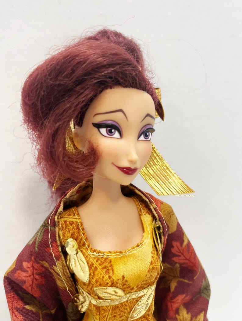 May include: A doll with reddish-brown hair styled in an updo, featuring purple eyeshadow and dark eyeliner. The doll wears a yellow bodice with gold leaf accents and a maroon outer garment patterned with brown and orange autumn leaves.