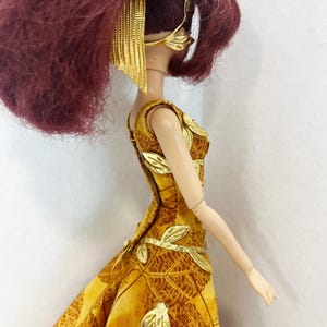 May include: A doll with dark red hair styled in a high ponytail, wearing a gold leaf-patterned headband and a sleeveless yellow and brown dress featuring a leaf print. The dress is adorned with metallic gold leaf appliqu&eacute;s along the bodice and skirt.