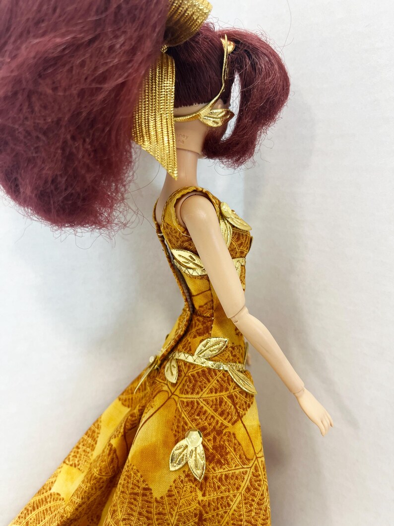 May include: A doll with dark red hair styled in a high ponytail, wearing a gold leaf-patterned headband and a sleeveless yellow and brown dress featuring a leaf print. The dress is adorned with metallic gold leaf appliqu&eacute;s along the bodice and skirt.