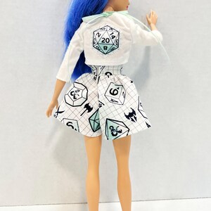May include: A doll with bright blue hair wearing a white long-sleeve cropped jacket featuring a 20-sided die graphic on the back and a white skirt with a black grid pattern and various gaming die icons in black and light blue colours.
