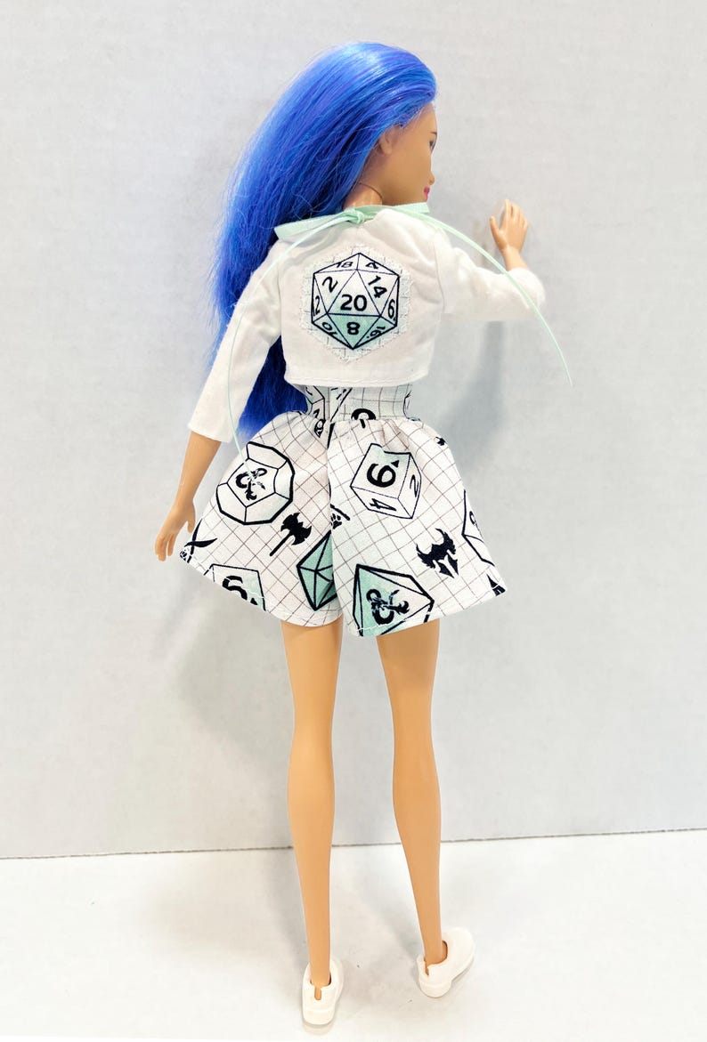 May include: A doll with bright blue hair wearing a white long-sleeve cropped jacket featuring a 20-sided die graphic on the back and a white skirt with a black grid pattern and various gaming die icons in black and light blue colours.